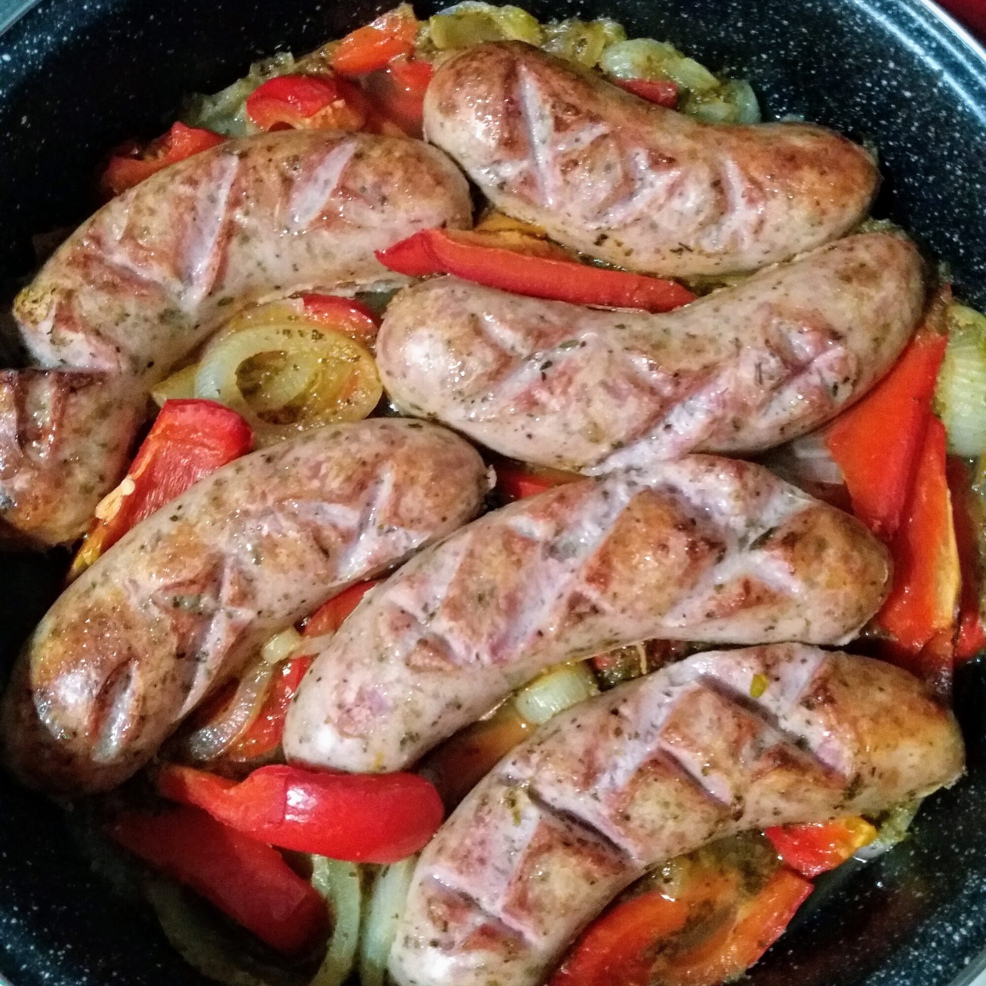 A pan of scored, white sausages with onion and red bell peppers.